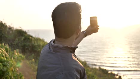 Ein-glücklicher-junger-Mann-oder-Reisenden-unter-ein-Bild-eines-Sonnenuntergangs-auf-sein-Handy-oder-Smartphone-der-untergehenden-Sonne.