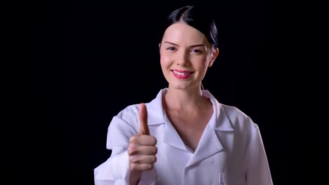 Confident-young-female-scientist-in-white-coat-showing-thumbs-up-into-camera-and-smiling,-isolated-on-black-background