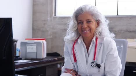 Senior-female-doctor-sitting-at-desk-in-an-office,-smiling