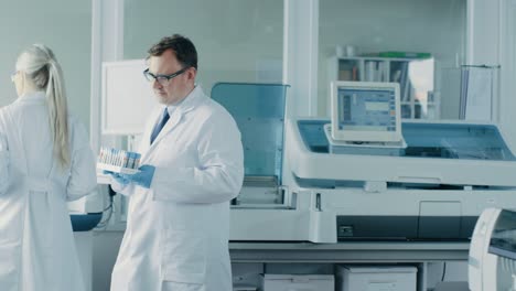 Male-Research-Scientist-Walks-Through-Laboratory-with-Tray-of-Test-Tubes-Filled-with-Samples.-In-the-Background-People-Working-in-Laboratory-with-Innovative-Equipment.-In-Slow-Motion.