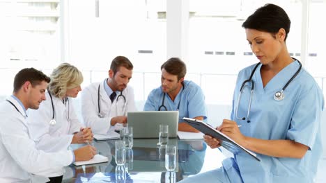 Thoughtful-surgeon-writing-on-clipboard-with-staff-talking-behind-her