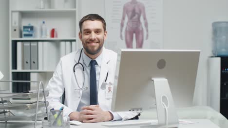 Male-Doctor-is-Working-at-His-Desk.-He-Interrupts-His-Work,-Folds-His-Hands-and-Smiles-at-the-Camera.