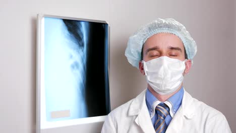 A-young-doctor-in-a-face-mask-looks-at-the-camera-and-smiles.-Protective-hat-on-the-head.-Against-the-background-hanging-x-ray-of-the-patient.-Shirt-with-a-tie-and-a-stethoscope-on-the-neck