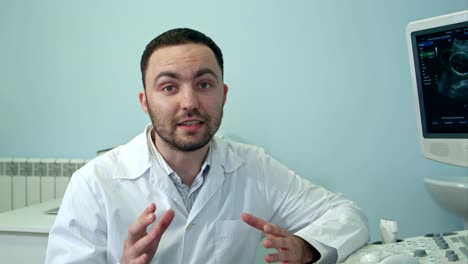 Young-male-doctor-talking-to-the-camera-sitting-next-to-ultrasound-scanning-machine