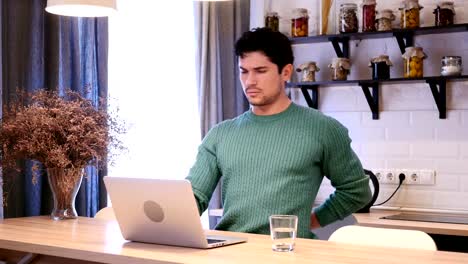 Young-Man-with-Back-Pain-Working-on-Laptop--in-Kitchen