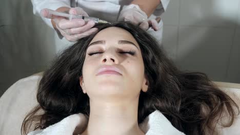 Handsome-woman-receives-an-injection-in-the-head.-The-procedure-makes-doctor-in-white-gloves.-The-concept-of-mesotherapy.-Thrust-to-strengthen-the-hair-and-their-growth