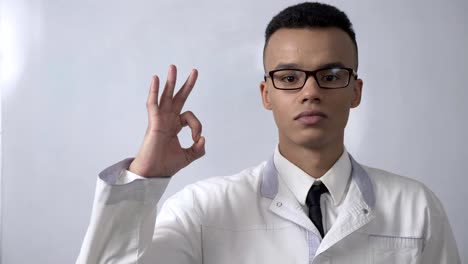Young-successful-African-scientist-in-a-white-coat-and-glasses-showing-ok-sign,-looks-at-the-camera,-portrait-concept.-60-fps