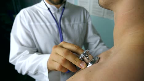 Portrait-of-handsome-doctor-examining-patient-with-stethoscope.-Young-medical-worker-listening-heartbeat-of-sick.-Caucasian-medic-checking-breast-of-unrecognizable-guy-in-his-office-at-the-hospital.-Close-up