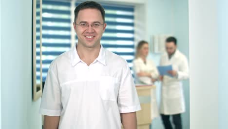 Sonriente-médico-masculino-en-gafas-mirando-a-cámara-mientras-personal-médico-que-trabaja-en-el-fondo