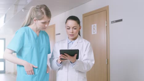 In-the-Hospital,-Busy-Doctors-Talk,-Using-Tablet-Computer-While-Standing-in-the-Hallway.-New-Modern-Fully-Functional-Medical-Facility.