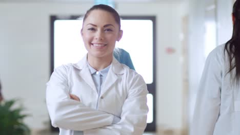 Schöne-junge-Frau-Doktor-Spaziergänge-durch-den-Krankenhaus-Flur,-She-Haltestellen,-kreuzt-Arme-und-charmant-Lächeln.-Professionelle-Leute-bei-der-Arbeit.