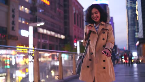 Zeitlupe-einer-ziemlich-glücklich-junge-asiatische-Frau-zu-Fuß-in-die-Stadt,-die-Straße-am-Abend-mit-Handy-in-der-hand