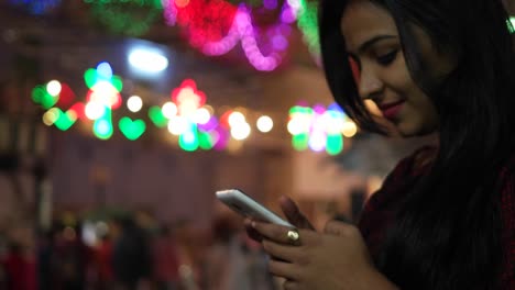 Joven-mujer-India-en-un-touch-pantalla-smart-teléfono-móvil-textos-mensajes-tipos-acciones-foto-video-llamadas-delante-de-un-brillante-colorido-festival-luces-fuera-de-foco-en-el-amor-de-fondo-celebración-mela