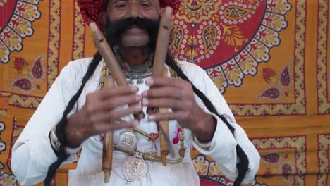 Hand-held-Rajasthani-elderly-male-starts-to-play-the-flute-with-his-nose-in-front-of-a-colourful-fabric-tent