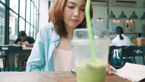 Cheerful-happy-asian-young-woman-sitting-drinking-iced-green-tea-in-cafe-using-smartphone-for-talking,-reading-and-texting.-Women-lifestyle-concept.