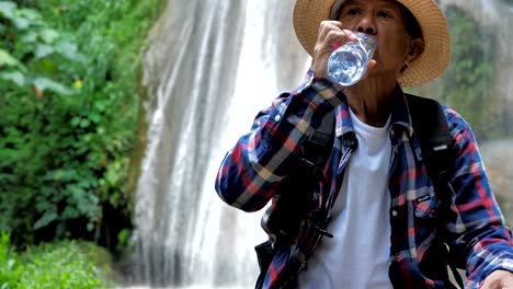 Close-Up-Senior-man-sitting-enjoying-by-a-waterfall-and-drink-Water.-Travel-Lifestyle-vacations-into-the-wild-wearing-cozy-shirt,hat-with-backpack.-Video-Slow-motion