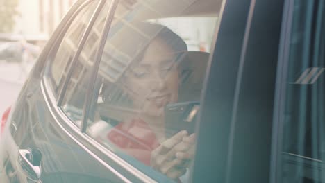 Schöne-Frau-in-einem-Auto-fährt,-sitzt-auf-einem-PKW-Rücksitz-nutzt-Smartphone,-browst-durch-Internet-Chats-mit-Freunden.-Kamera-Schuss-von-außerhalb-des-Fahrzeugs.