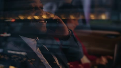 Handsome-Businessman-and-Beautiful-Businesswoman-Riding-on-the-Backseat-of-a-Car-in-the-Evening.-Man-Makes-a-Phone-Call,-Woman-Works-on-a-Laptop.-Camera-Shot-made-from-Outside-the-Car.