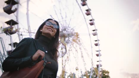 Mujer-joven-con-estilo-en-gafas-sonriendo-alegremente-delante-de-una-noria-en-el-parque-de-atracciones