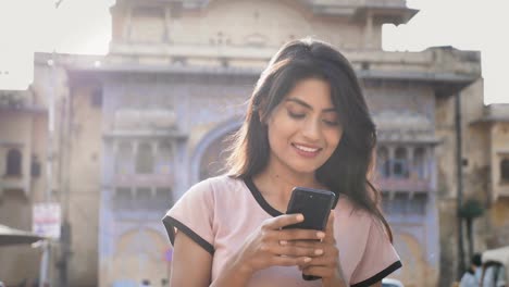 Una-mujer-joven-y-atractiva-en-una-ciudad-calle-y-utilizando-smartphone-o-teléfono-móvil-con-una-sonrisa.