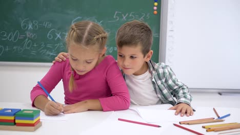 school-love,-classmates-write-during-lesson-at-table-and-then-look-at-camera-and-smile-on-background-of-blackboard