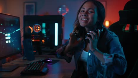 Portrait-of-a-Pretty-and-Excited-Black-Gamer-Girl-in-Headphones-Playing-First-Person-Shooter-Online-Video-Game-on-Her-Computer.-She-Turns-and-Smiles-into-the-Camera.-Home-is-Lit-with-Neon-Lights.