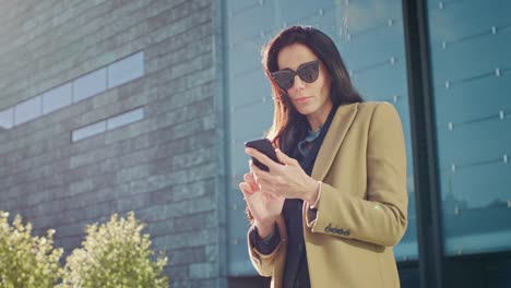 Elegante-empresaria-usa-Smartphone-para-realizar-negocios-mientras-pie-exterior-moderno-edificio-de-cristal.-Hermosa-mujer-elegante-vestida-con-abrigo-y-gafas-oscuras-paseos-en-entorno-urbano-de-la-ciudad-moderna.