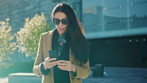 Elegante-empresaria-usa-Smartphone-para-realizar-negocios-mientras-camina-por-el-edificio-de-cristal-moderno.-Hermosa-mujer-elegante-vestida-con-abrigo-y-gafas-oscuras-paseos-en-entorno-urbano-de-la-ciudad-moderna.