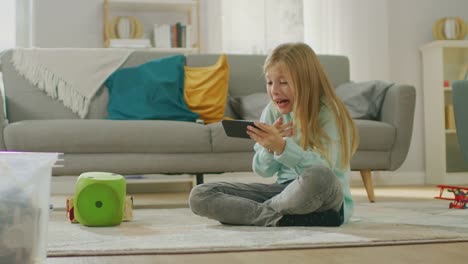 Smart-Cute-Girl-Sitting-on-a-Carpet-Playing-in-Video-Game-on-His-Smartphone,-Holds-and-Uses-Mobile-Phone-in-Horizontal-Landscape-Mode.-Child-Has-Fun-Playing-Videogame-in-Sunny-Living-Room.