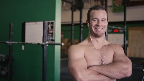 Ajuste-Retrato-de-un-hombre-joven-con-auriculares-sonriendo-y-cruzando-sus-brazos-en-un-pequeño-gimnasio