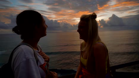 Two-happy-female-travelers-talking-and-laughing-at-the-sunset-against-a-background-of-the-sea.-Blonde-and-brunette-are-chattering-and-smiling-looking-at-waving-ocean-behind-them