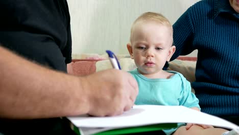 Attraktive-junge-zieht-einen-Stift-mit-seinen-Großeltern-nach-Hause-auf-der-Couch.-Der-junge-starrt-auf-die-Tiere,-die-Großvater-zog.-Das-Konzept-der-verschiedenen-Generationen