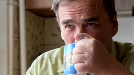 An-elderly-man-with-a-mustache-holds-a-cup-of-hot-drink-and-drinks.-He-sits-near-the-window-and-has-breakfast-at-home