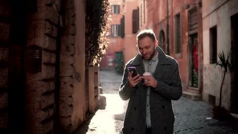 Retrato-de-joven-feliz-caminar-en-la-ciudad-y-el-uso-de-smartphone.-Hombre-navegar-Internet-y-explotación-taza-de-café