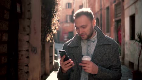 Portrait-of-Handsome-man-standing-in-the-little-street-and-using-touchscreen-technology-at-smartphone