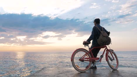 Joven-mujer-sentada-en-la-bicicleta-vintage-en-el-paseo-marítimo-durante-el-hermoso-amanecer