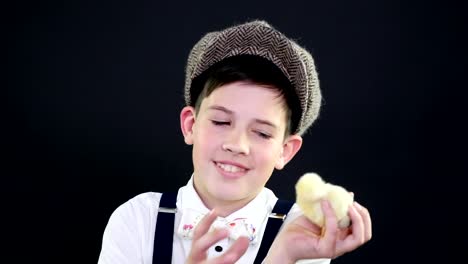 Portrait,-a-pretty-boy-in-a-cap-and-suspenders-plays-with-a-small-yellow-duckling.-Studio-video-with-thematic-decoration