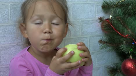 Niedliche-kleine-Mädchen-sitzen-auf-dem-Boden-und-Essen-Apfel-neben-einem-Weihnachtsbaum.