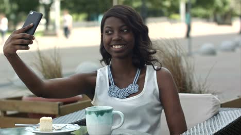 Attractive-young-african-woman-smiling-and-taking-a-selfie-with-her-smartphone-while-sitting-alone-in-outdoor-cafe-enjoying-a-meal