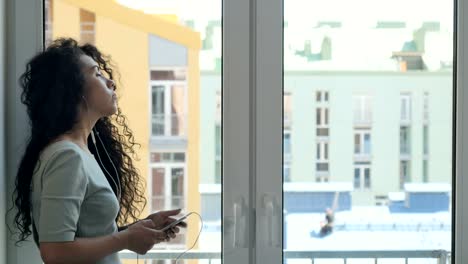 Curly-brunette-messaging-with-someone-listening-a-music-near-the-window