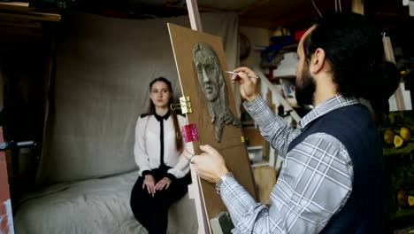 Skilled-sculptor-works-with-plasticine-on-canvas-to-create-woman's-face-of-posing-model-in-art-studio