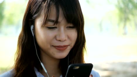 Cute-woman-is-reading-pleasant-text-message-on-mobile-phone-while-sitting-in-the-park-in-warm-spring-day,gorgeous-female-is-listening-to-music-in-headphones-and-searching-information-on-cell-telephone