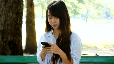 Cute-woman-is-reading-pleasant-text-message-on-mobile-phone-while-sitting-in-the-park-in-warm-spring-day,gorgeous-female-is-listening-to-music-in-headphones-and-searching-information-on-cell-telephone