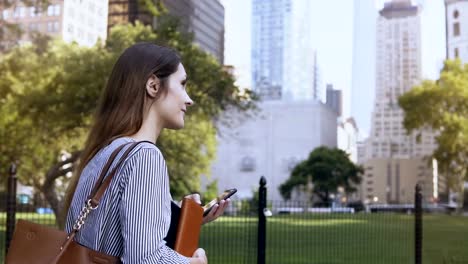 Junge-schöne-Frau-zu-Fuß-durch-den-Park-bei-der-Arbeit.-Geschäftsfrau-hält-die-Dokumente-und-mit-smartphone