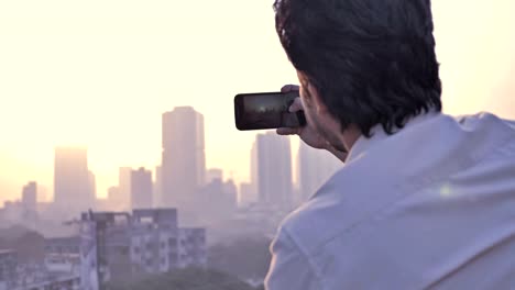 Un-hombre-joven-clic-en-fotos-de-una-puesta-de-sol-sobre-el-horizonte-de-una-ciudad-usando-un-teléfono-celular