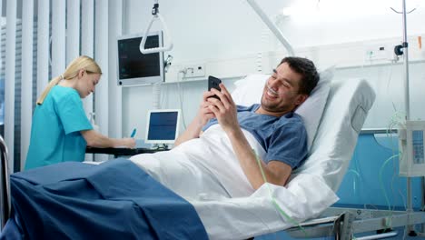 Recovering-Patient-Uses-Smartphone-and-Smiles-while-Lying-on-a-Bed-in-the-Hospital.-Friendly-Nurse-Comes-in-to-Check-on-Him.