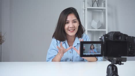 Glücklich-lächelnd-asiatische-Frau-oder-Beauty-Blogger-mit-Pinsel-und-Kamera-Videoaufnahme-und-winkenden-Hand-zu-Hause.-Videoblog-Blogging-Menschen-Schönheitskonzept.-Dolly-erschossen.