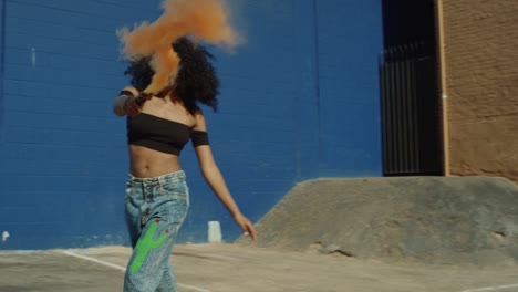 Beautiful-young-woman-holding-colorful-smoke-grenade-dancing-outside-against-blue-wall