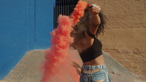 Beautiful-young-woman-holding-colorful-smoke-grenade-dancing-outside-against-blue-wall