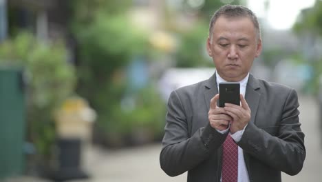 Reife-japanischer-Geschäftsmann-mit-Telefon-in-den-Straßen-im-freien